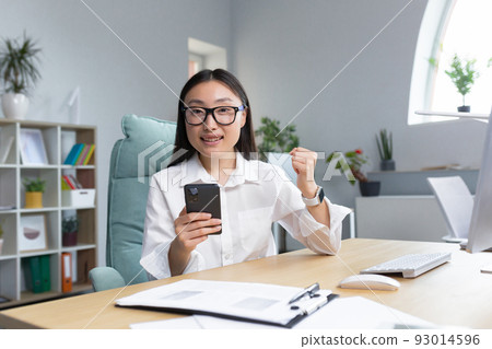 Young beautiful Asian businesswoman working in office, smiling and happy with victory, using phone, reading good news, celebrating triumph. 93014596