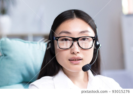 Close-up portrait of a young beautiful asian woman with glasses smiling and looking at the camera, uses a headset for video communication call center employee. Close-up portrait of a young beautiful asian woman with glasses smiling and looking at the camera, uses a headset for video communication call center employee. 93015276