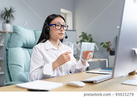 Consultant woman working remotely using headset for video call, talking to customers, helpline, Asian woman working in office. 93015279