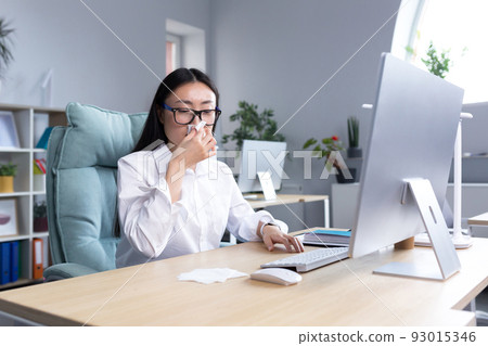 Sick Asian female office worker, has cold and flu, coughs and sneezes into tissue, works at computer. Sick Asian female office worker, has cold and flu, coughs and sneezes into tissue, works at computer. 93015346