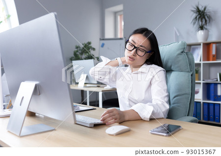 Overtired Asian woman office worker, has severe neck pain, massages neck muscles with hand, female worker in office at computer. Overtired Asian woman office worker, has severe neck pain, massages neck muscles with hand, female worker in office at computer. 93015367