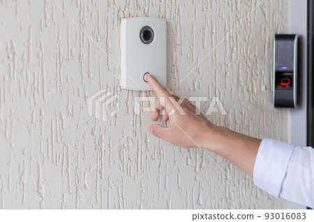 Hand of a man in a white shirt pressing the button of a door knocker on a white wall of a house, a smart door knocker with a video camera for communication 93016083