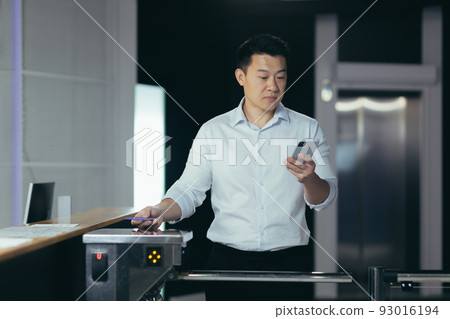 Successful Asian businessman worker enters office, uses key card to open turnstile, man looks at phone screen reads news Successful Asian businessman worker enters office, uses key card to open turnstile, man looks at phone screen reads news 93016194