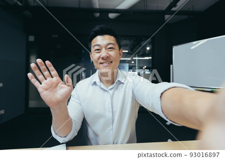 Asian businessman working in modern office, successful man talking on video phone call, waving to interlocutor and smiling Asian businessman working in modern office, successful man talking on video phone call, waving to interlocutor and smiling 93016897