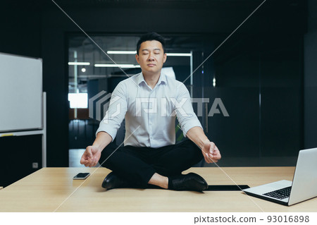 Asian boss businessman meditating in office, man sitting in lotus position on desk with closed eyes, thinking about important decision 93016898