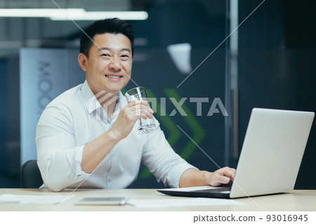 Asian businessman in the office drinking clean filtered water, portrait of a man smiling and looking at the camera holding a glass of water, a man at work with a laptop Asian businessman in the office drinking clean filtered water, portrait of a man smiling and looking at the camera holding a glass of water, a man at work with a laptop 93016945