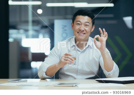 Portrait of successful asian businessman, man looking at camera and smiling showing ok sign, holding glass of clean water Portrait of successful asian businessman, man looking at camera and smiling showing ok sign, holding glass of clean water 93016969