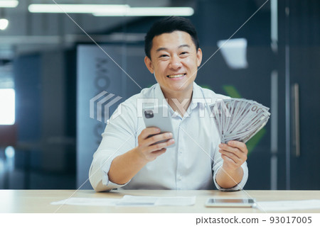 Successful asian businessman working in office, man smiling and looking at camera, holding phone and money dollars cash Successful asian businessman working in office, man smiling and looking at camera, holding phone and money dollars cash 93017005