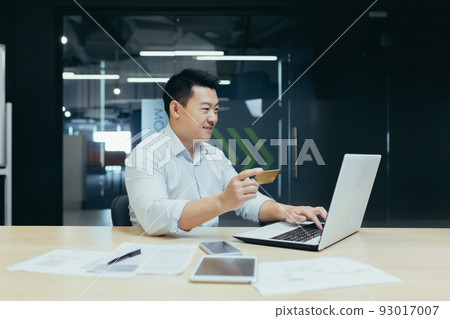 Cheerful and smiling asian businessman working in modern office, dark colors, making online purchase, using bank credit card and laptop, for online shopping Cheerful and smiling asian businessman working in modern office, dark colors, making online purchase, using bank credit card and laptop, for online shopping 93017007