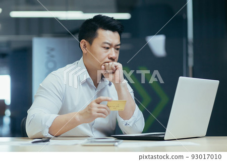 Thoughtful and serious Asian businessman trying to make a bank transaction, man working in office with laptop, holding credit bank card 93017010