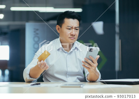 Frustrated Asian businessman with credit card and phone, working in office, trying to make online purchase, and bank transfer 93017013