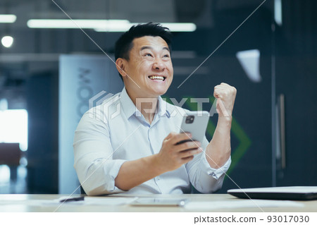 Happy and successful Asian businessman working in office, typing on phone and reading good news, celebrating triumph and victory 93017030
