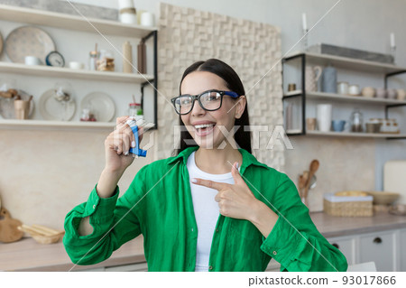 Young beautiful woman in glasses and green shirt at home holds asthma and allergy inhaler in hand, shows it with finger of other hand, advertises, recommends, smiles 93017866