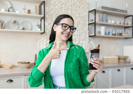 Young satisfied woman at home eating chocolate bar in kitchen, brunette in glasses and green shirt holds chocolate in hands and points to it with finger 93017988
