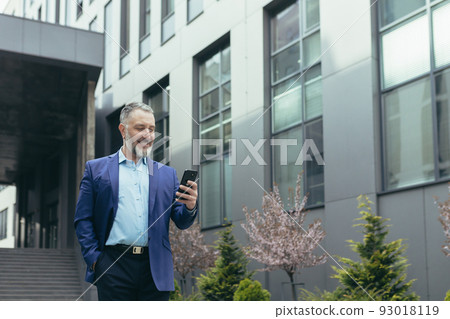 Senior experienced gray-haired businessman outside office, smiling and happy using phone, boss reading and looking at smartphone screen, walking near building 93018119