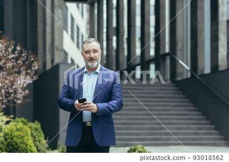 Successful senior investor with phone outside office building, businessman in business suit using smartphone and looking away dreamily Successful senior investor with phone outside office building, businessman in business suit using smartphone and looking away dreamily 93018562
