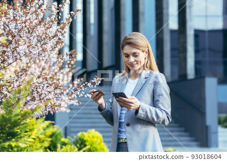 Beautiful blonde woman near a tree with flowers holds a phone and a bank credit card, smiles and makes purchases in the online store Beautiful blonde woman near a tree with flowers holds a phone and a bank credit card, smiles and makes purchases in the online store 93018604