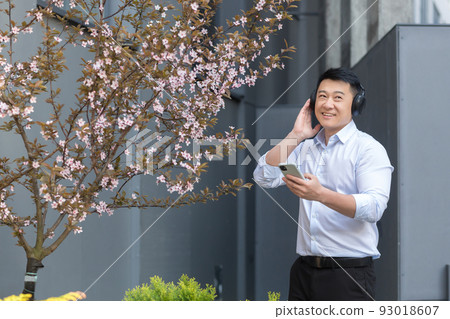 Cheerful and smiling successful Asian business man listening to music using smartphone and big headphones, manager resting outside office in shirt 93018607