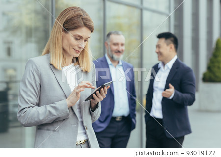 A beautiful and successful business woman is using the phone outside the office, on the photo are male colleagues talking, a successful business team is on a break A beautiful and successful business woman is using the phone outside the office, on the photo are male colleagues talking, a successful business team is on a break 93019172