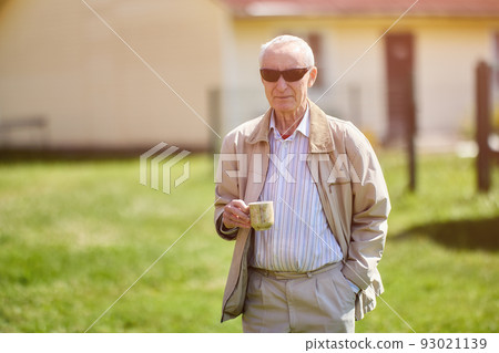 Serious elderly man 75 years old standing outdoors in front of his house with mug of hot drink in his hand and sunglasses on his face. 93021139