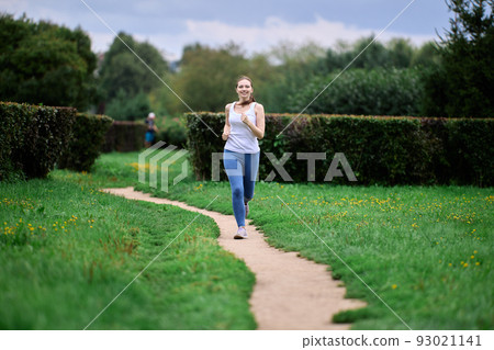Female jogger in white t-shirt singlet and leggings takes morning run in residential district of city next to multistorey apartment building. 93021141