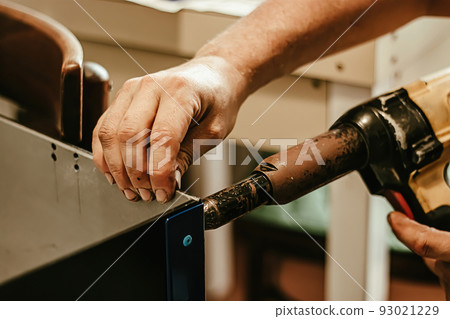 Male worker uses electric riveting gun. Hand holding riveting machine and placing rivets. Working workshop of carpenter or mechanic. 93021229