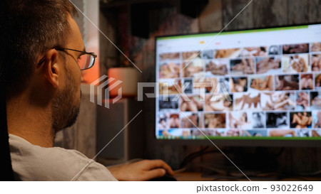 A bearded man with glasses looks at erotic pictures on the monitor at night. A close-up of his head. Sex addiction. Sex alone. A bearded man with glasses looks at erotic pictures on the monitor at night. A close-up of his head. Sex addiction. Sex alone. 93022649
