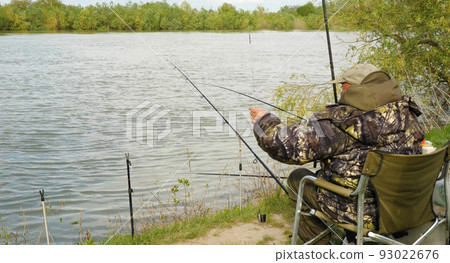 A fisherman with a fishing rod sits in an armchair on the bank of a river. There are green trees in the background. The concept of recreation and hobbies in nature. 93022676