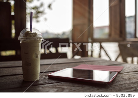 A glass with smoothies and a tablet on a wooden table on the veranda of a cafe. A glass with smoothies and a tablet on a wooden table on the veranda of a cafe. 93023899