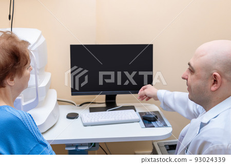 A male ophthalmologist explains the results of an eye examination to an adult woman, showing a drawing on a computer 93024339