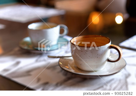 Orange and blue cups on saucers. The mugs with tea in soft focus standing on menu papers on naturally blurred background. Coffee, tea house, bokeh lights. The concept of a cozy pastime 93025282
