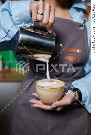 Top view shot of professional barista pouring milk from jar in to a cup of coffee, coffee being prepared by a barista. Focus on hands holding cup of coffee. Top view shot of professional barista pouring milk from jar in to a cup of coffee, coffee being prepared by a barista. Focus on hands holding cup of coffee. 93025630