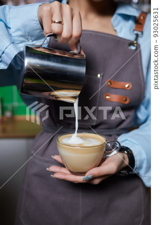 Top view shot of professional barista pouring milk from jar in to a cup of coffee, coffee being prepared by a barista. Focus on hands holding cup of coffee. Top view shot of professional barista pouring milk from jar in to a cup of coffee, coffee being prepared by a barista. Focus on hands holding cup of coffee. 93025631