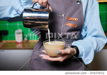 Top view shot of professional barista pouring milk from jar in to a cup of coffee, coffee being prepared by a barista. Focus on hands holding cup of coffee. 93025632
