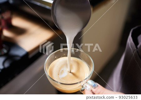 Top view shot of professional barista pouring milk from jar in to a cup of coffee, coffee being prepared by a barista. Focus on hands holding cup of coffee. Top view shot of professional barista pouring milk from jar in to a cup of coffee, coffee being prepared by a barista. Focus on hands holding cup of coffee. 93025633