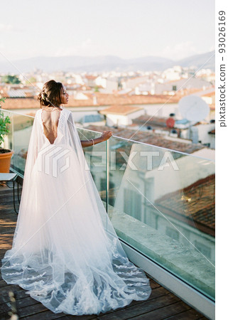 Bride stands near the glass railing and looks at the roofs of old houses Bride stands near the glass railing and looks at the roofs of old houses 93026169