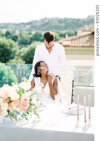 Groom hugs from behind bride sitting at the table on the terrace of the building Groom hugs from behind bride sitting at the table on the terrace of the building 93026305