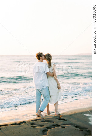Man hugs and kisses woman on the temple while standing on the beach. Back view Man hugs and kisses woman on the temple while standing on the beach. Back view 93026306