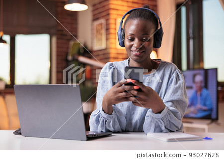 Smiling african american remote worker in headphones typing message on mobile phone. Female freelancer watching videos on smartphone in modern home office with beautiful warm sunset light Smiling african american remote worker in headphones typing message on mobile phone. Female freelancer watching videos on smartphone in modern home office with beautiful warm sunset light 93027628