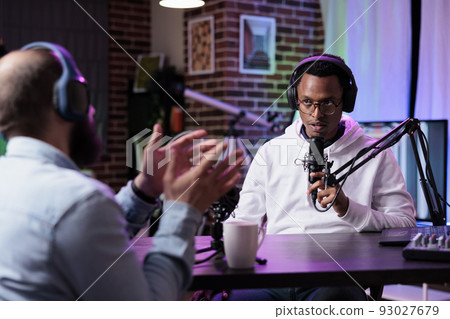 African american influencer recording podcast with guest, using sound equipment at desk. Cheerful people live broadcasting discussion for social media web, streaming online content. 93027679