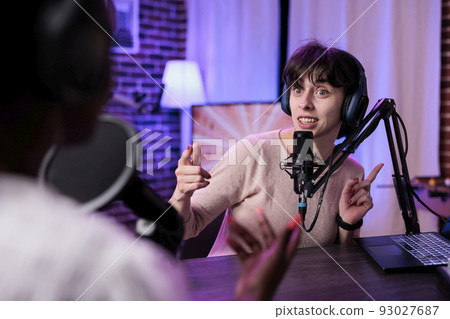 Female presenter interviewing young woman with podcast equipment, having conversation to broadcast live on channel. Women meeting in studio to record discussion on social media. 93027687