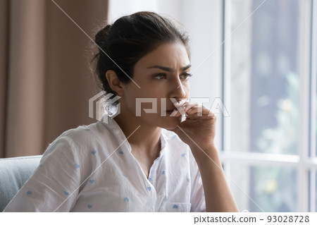 Serious thoughtful beautiful Indian woman sitting on couch indoors Serious thoughtful beautiful Indian woman sitting on couch indoors 93028728