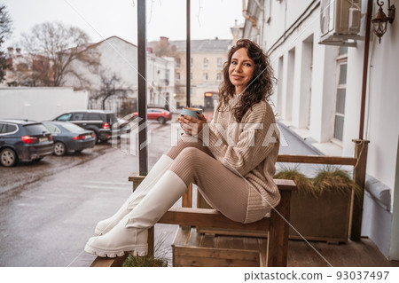 A middle-aged woman in a beige sweater with a blue mug in her hands is in a street cafe on the veranda A middle-aged woman in a beige sweater with a blue mug in her hands is in a street cafe on the veranda 93037497