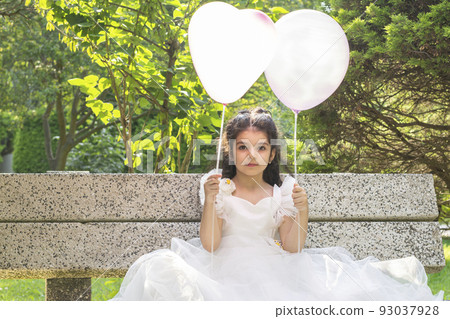 Little girl holding two balloons at hand seated on park bench 93037928