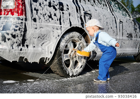 Toddler child leaning over rear wheel of foamy car and holding sponge for washing auto, testing himself in the profession of car washer outdoors in the afternoon. 93038416