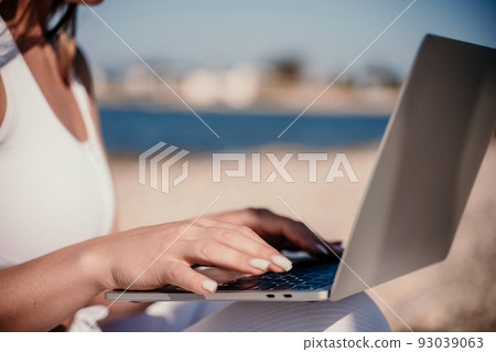 young beautiful woman with dark hair in white overalls sits on the sand near the sea and uses a laptop 93039063