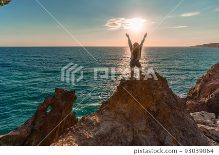 A female tourist, standing with her hands up, reaches the ends of the earth and sees the dragon rock. The girl welcomes the sun. Concept design. A successful woman looks at the sunset with open arms. A female tourist, standing with her hands up, reaches the ends of the earth and sees the dragon rock. The girl welcomes the sun. Concept design. A successful woman looks at the sunset with open arms. 93039064