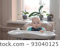 hungry boy sits waiting for food at an empty table for feeding in the kitchen on the windowsill of an orchid. 93040125