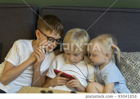 Three children sit on couch and look at the phone. Siblings enthusiastically look at screen of smartphone. Modern children. 93041858