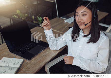 Young woman using headset and computer while talking with customers online Young woman using headset and computer while talking with customers online 93044408
